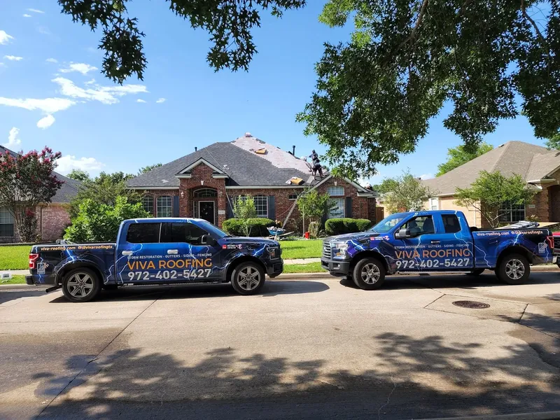 Two blue Viva Roofing trucks parked in front of a brick home while the crew works on the roof